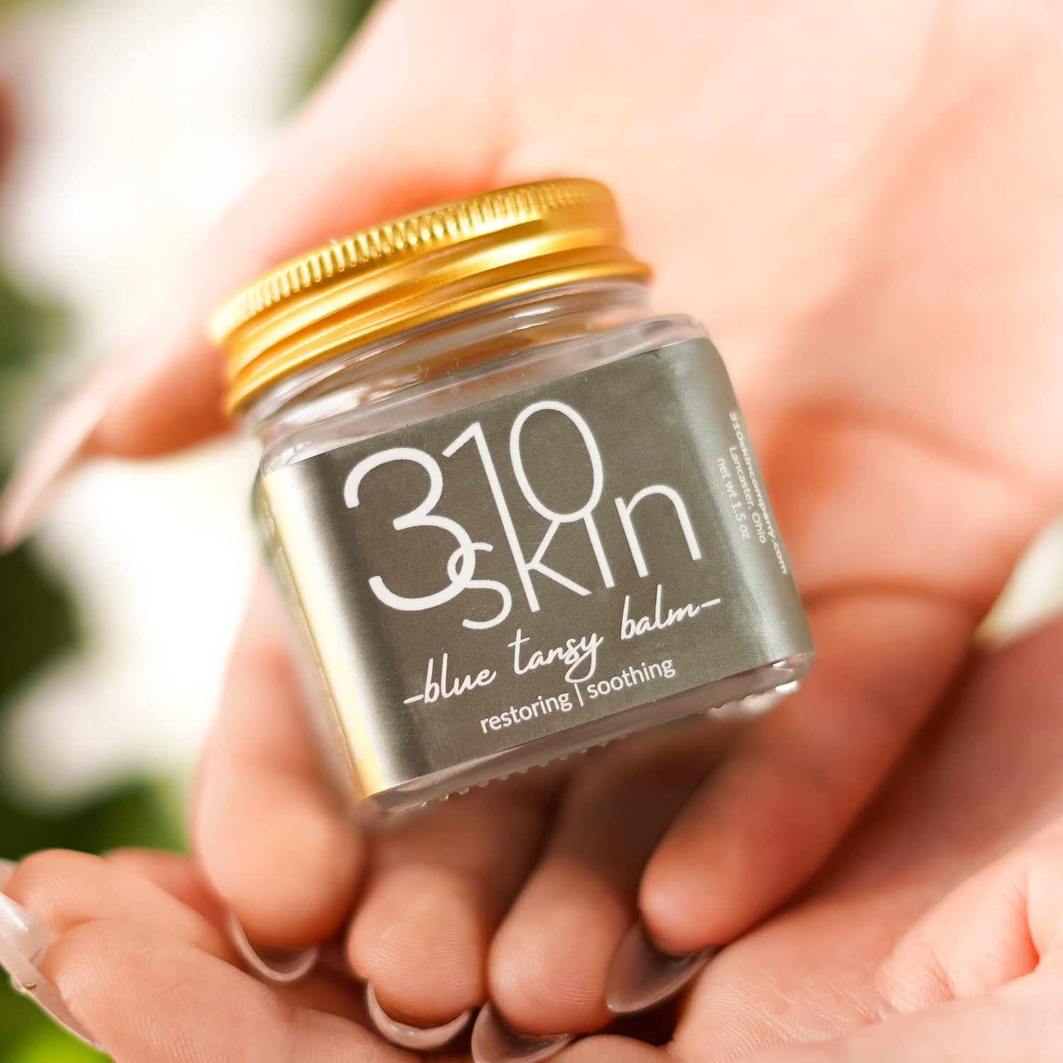 Hand holding a small jar of 310 Skin blue tansy balm with a blurred green background